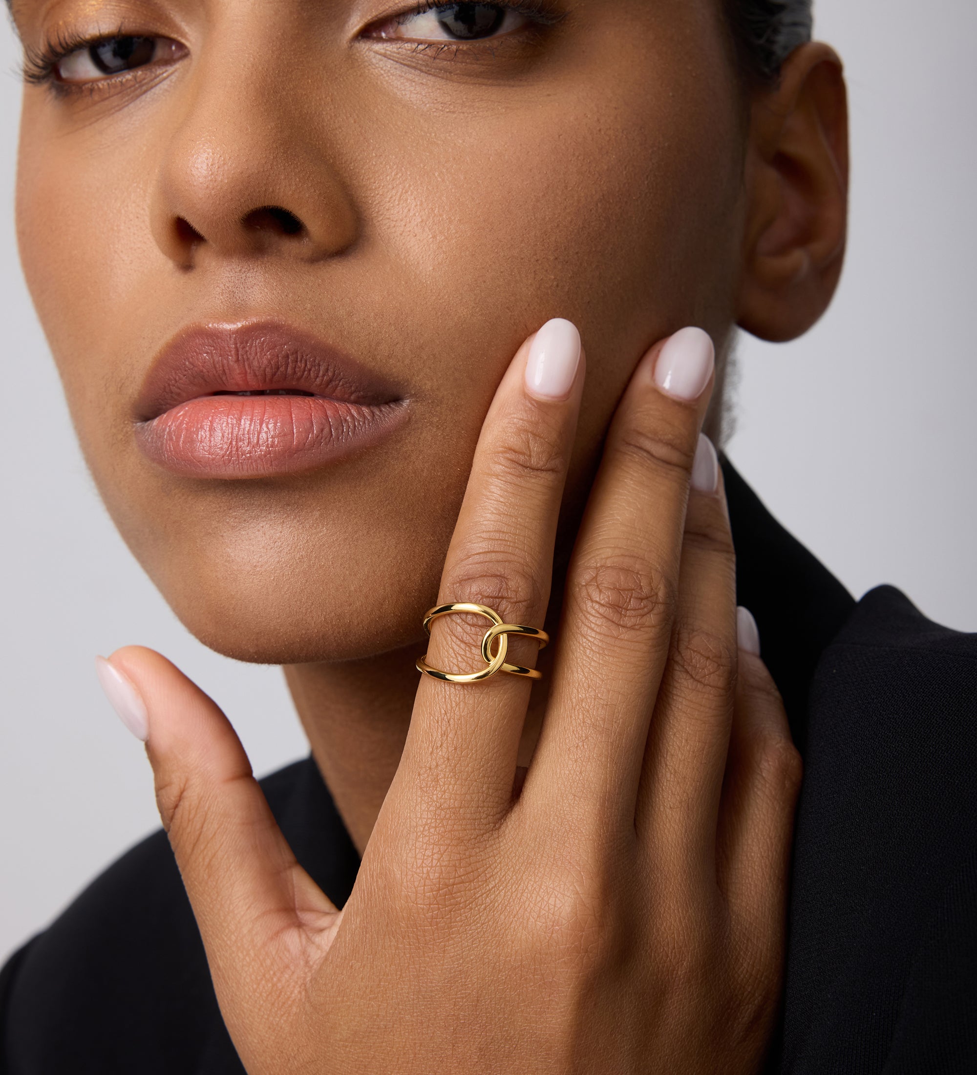 A person with smooth brown skin and nude nails touches their face, showcasing the Ayla Link Ring in Gold on their finger. They wear a black outfit and have a calm, confident expression.