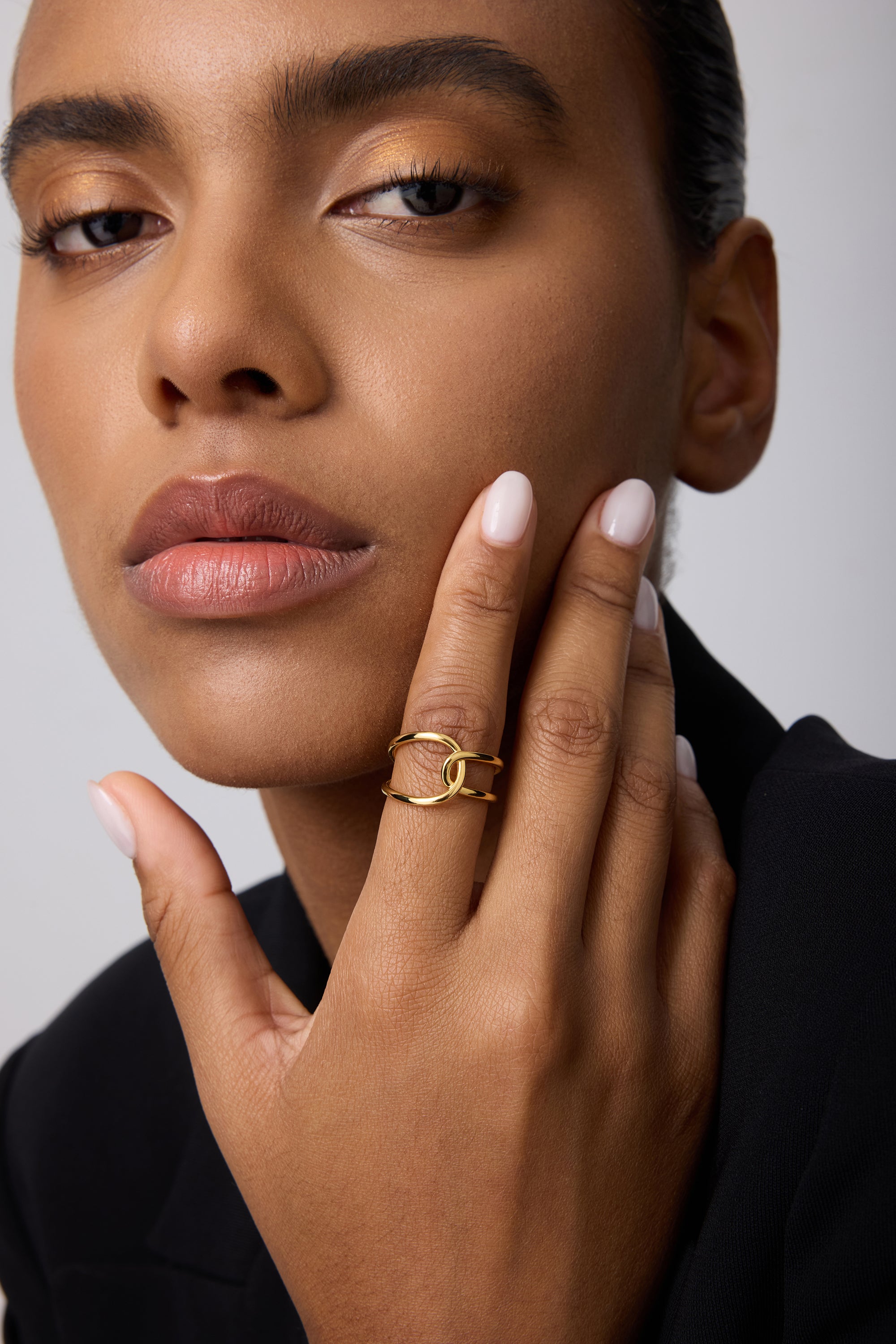 A person with smooth brown skin and nude nails touches their face, showcasing the Ayla Link Ring in Gold on their finger. They wear a black outfit and have a calm, confident expression.