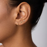 Close-up of a woman's ear with multiple piercings, featuring 14K Recycled Solid Yellow Gold Twisted Huggie Earrings among other sustainable gold and silver styles against a neutral background.