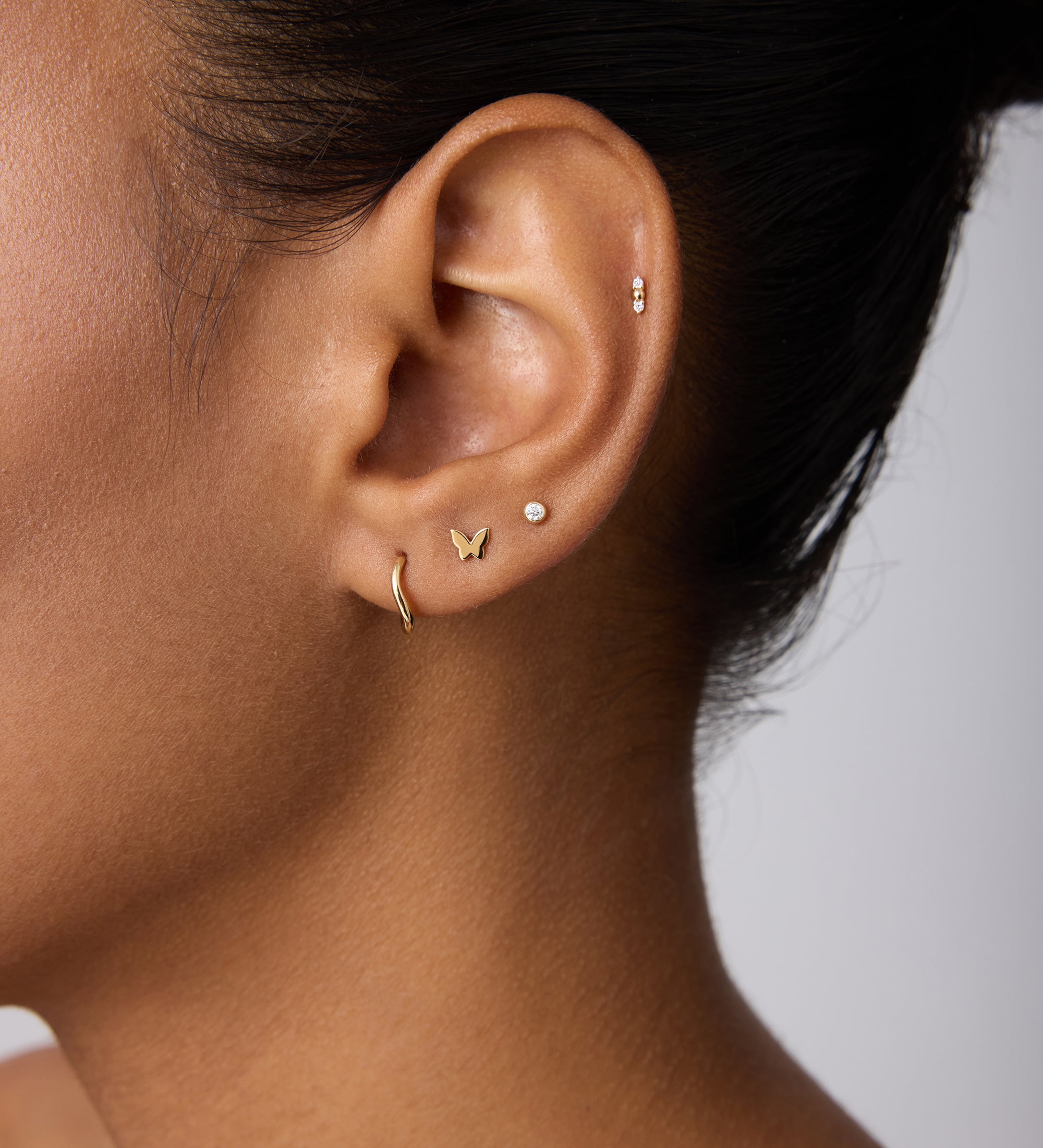 Close-up of a woman's ear with multiple piercings, featuring 14K Recycled Solid Yellow Gold Twisted Huggie Earrings among other sustainable gold and silver styles against a neutral background.