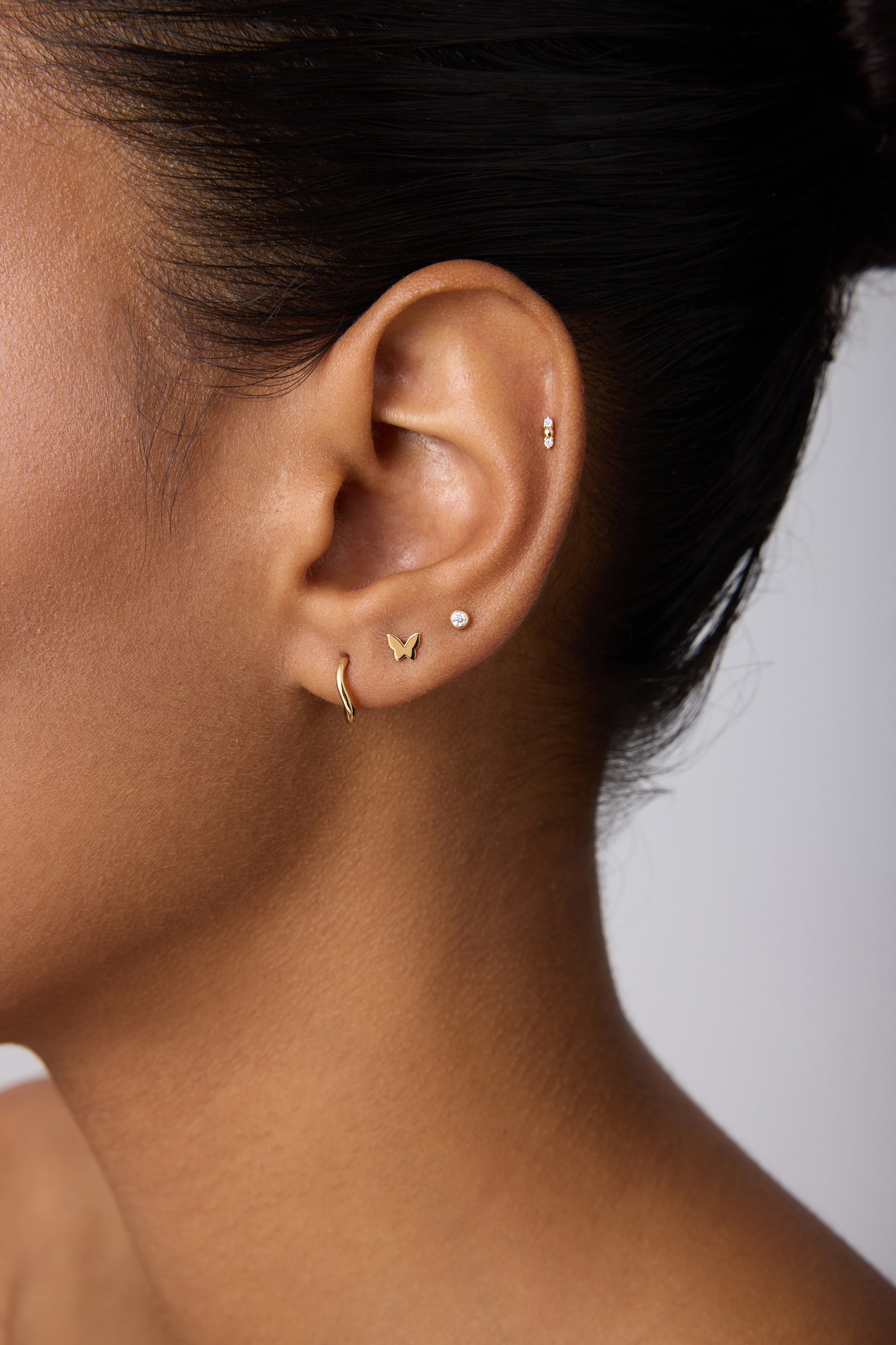 Close-up of a woman's ear with multiple piercings, featuring 14K Recycled Solid Yellow Gold Twisted Huggie Earrings among other sustainable gold and silver styles against a neutral background.