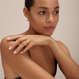 A woman with smooth, medium skin and slicked-back hair rests her chin on her hand, wearing gold hoop earrings, a thin bracelet, the Yvette Two-Tone Mixed Metal Ring and another ring, plus a black top, natural makeup and light pink nails.