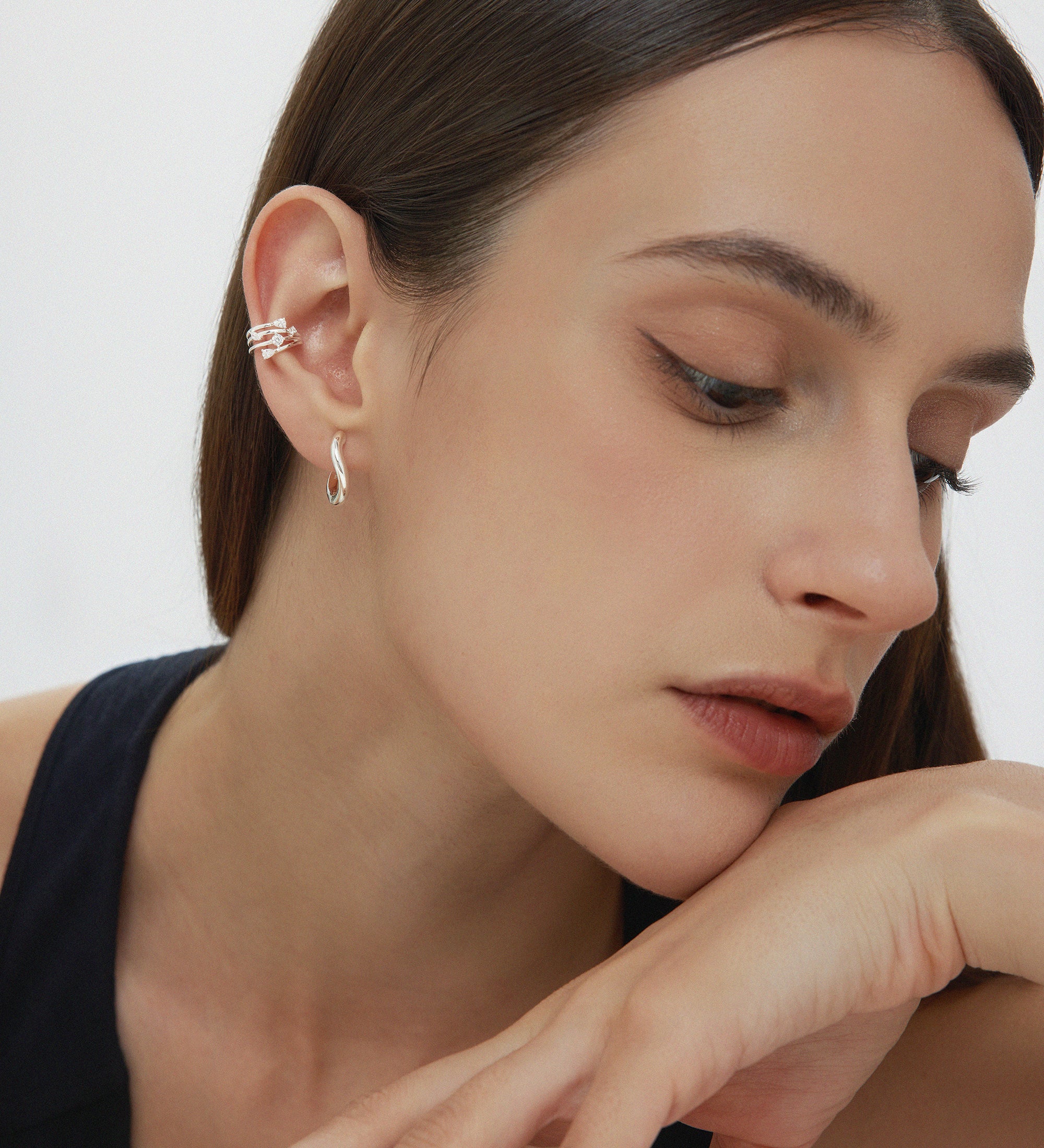 A woman with straight brown hair wears the Vale Crystal Ear Cuff in Silver and a drop earring, resting her chin on her hand. She has natural makeup and a black top, posed against a plain white background.