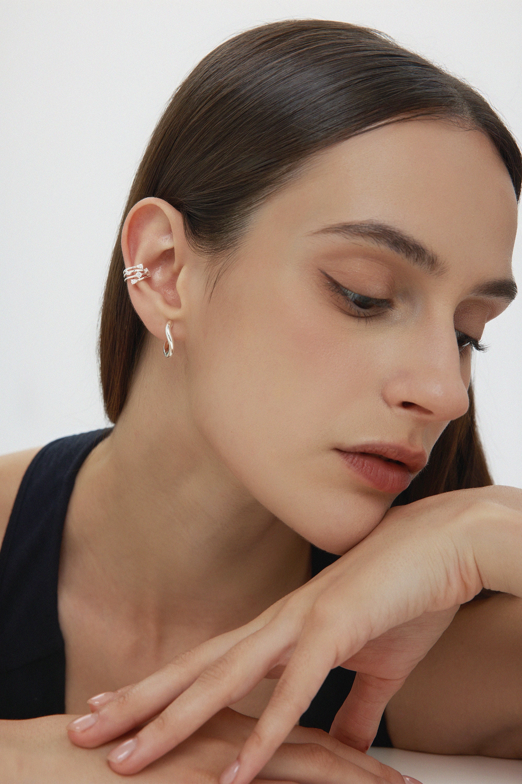 A woman with straight brown hair wears the Vale Crystal Ear Cuff in Silver and a drop earring, resting her chin on her hand. She has natural makeup and a black top, posed against a plain white background.