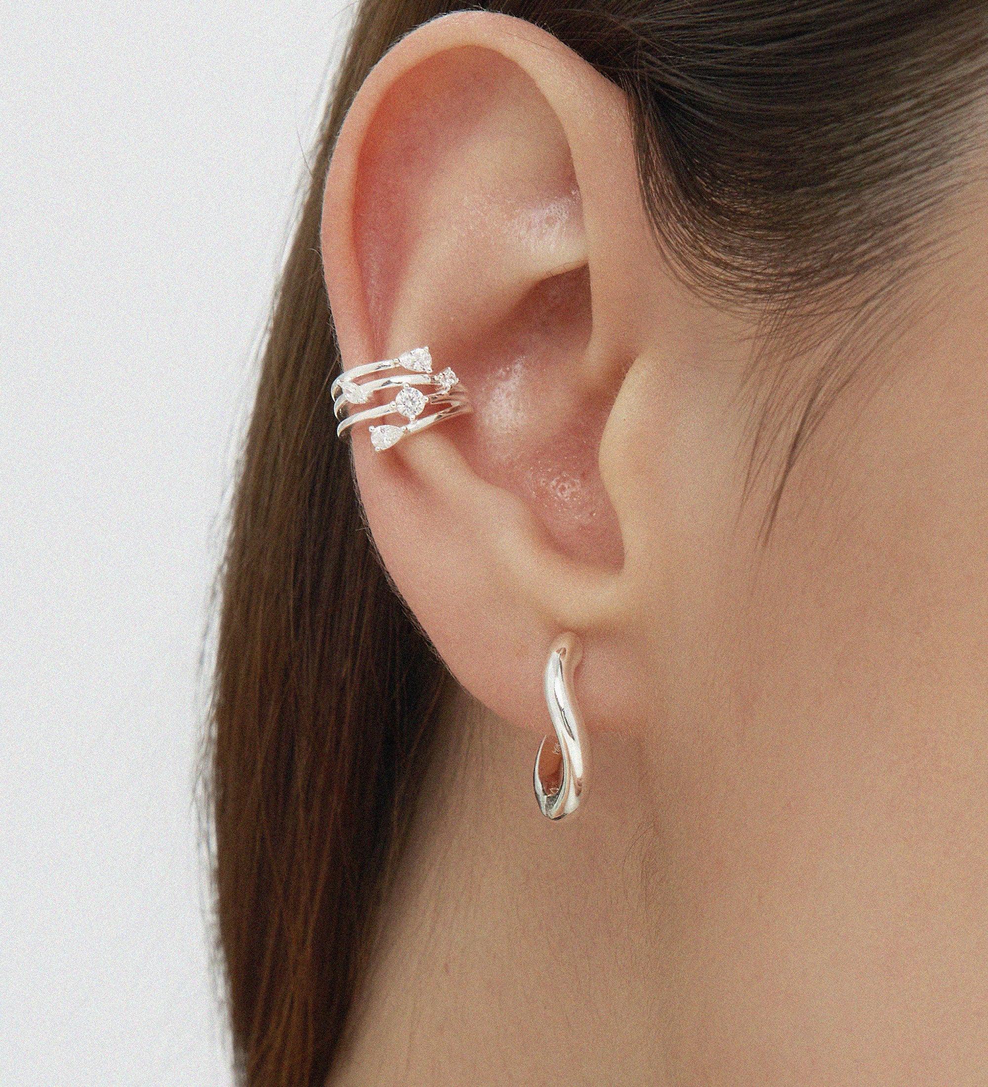 A close-up shows brown hair tucked behind an ear styled with an elegant stack: the Vale Crystal Ear Cuff in Silver featuring sparkling gemstones on the upper ear and a twisted silver hoop earring in the lobe.