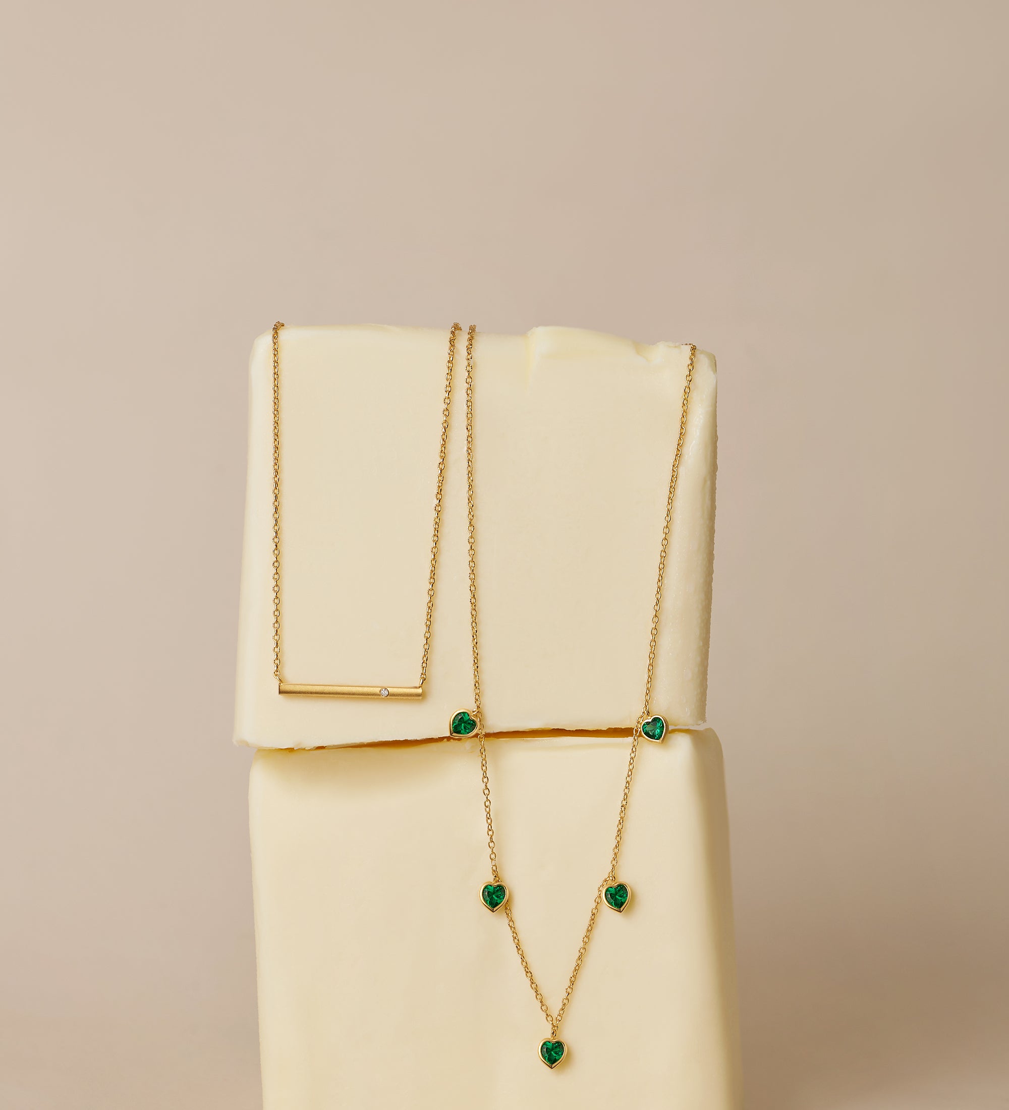 The Thea Green Heart Pendant Gold Necklace, featuring a small green cubic zirconia heart, is draped over two stacked blocks of butter against a neutral beige background.