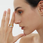 A woman with dark hair pulled back showcases the Vava Plain Chunky Curved Wave Ring in Gold, stacked with a minimalist gold-plated ring. She poses with a neutral expression against a plain background, her shoulders bare.