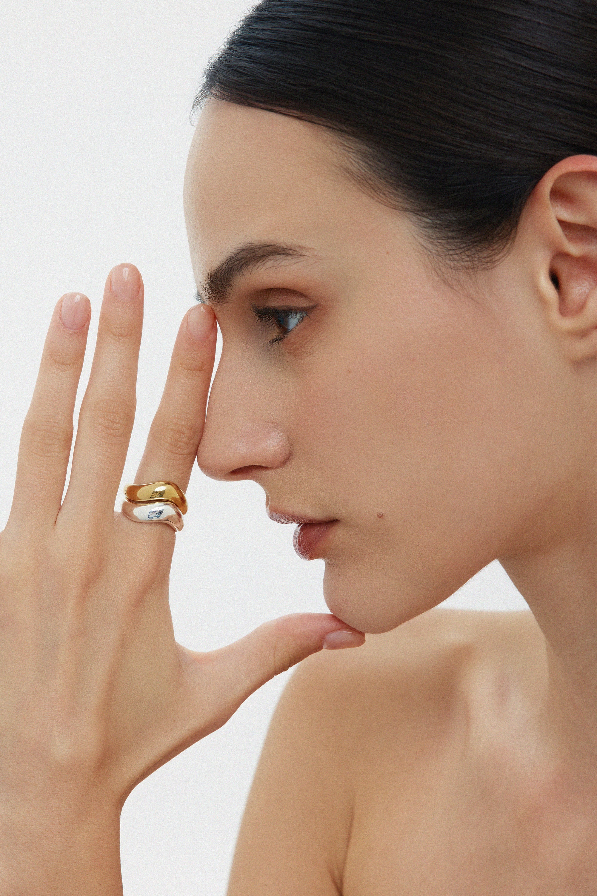 A woman with dark hair pulled back showcases the Vava Plain Chunky Curved Wave Ring in Gold, stacked with a minimalist gold-plated ring. She poses with a neutral expression against a plain background, her shoulders bare.