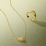 Displayed on a light green textured surface, the Vava Plain Chunky Curved Wave Ring in Gold and a matching abstract necklace cast soft shadows—a striking minimalist jewelry duo.