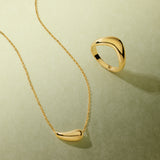 Displayed on a light green textured surface, the Vava Plain Chunky Curved Wave Ring in Gold and a matching abstract necklace cast soft shadows—a striking minimalist jewelry duo.