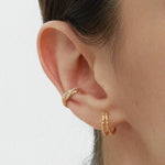 A close-up of a person’s ear wearing two gold hoop earrings and the Vera Twisted Crystal Ear Cuff in Gold, all against a plain light background.