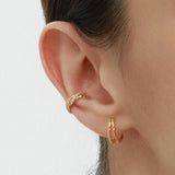 A close-up of a person’s ear wearing two gold hoop earrings and the Vera Twisted Crystal Ear Cuff in Gold, all against a plain light background.