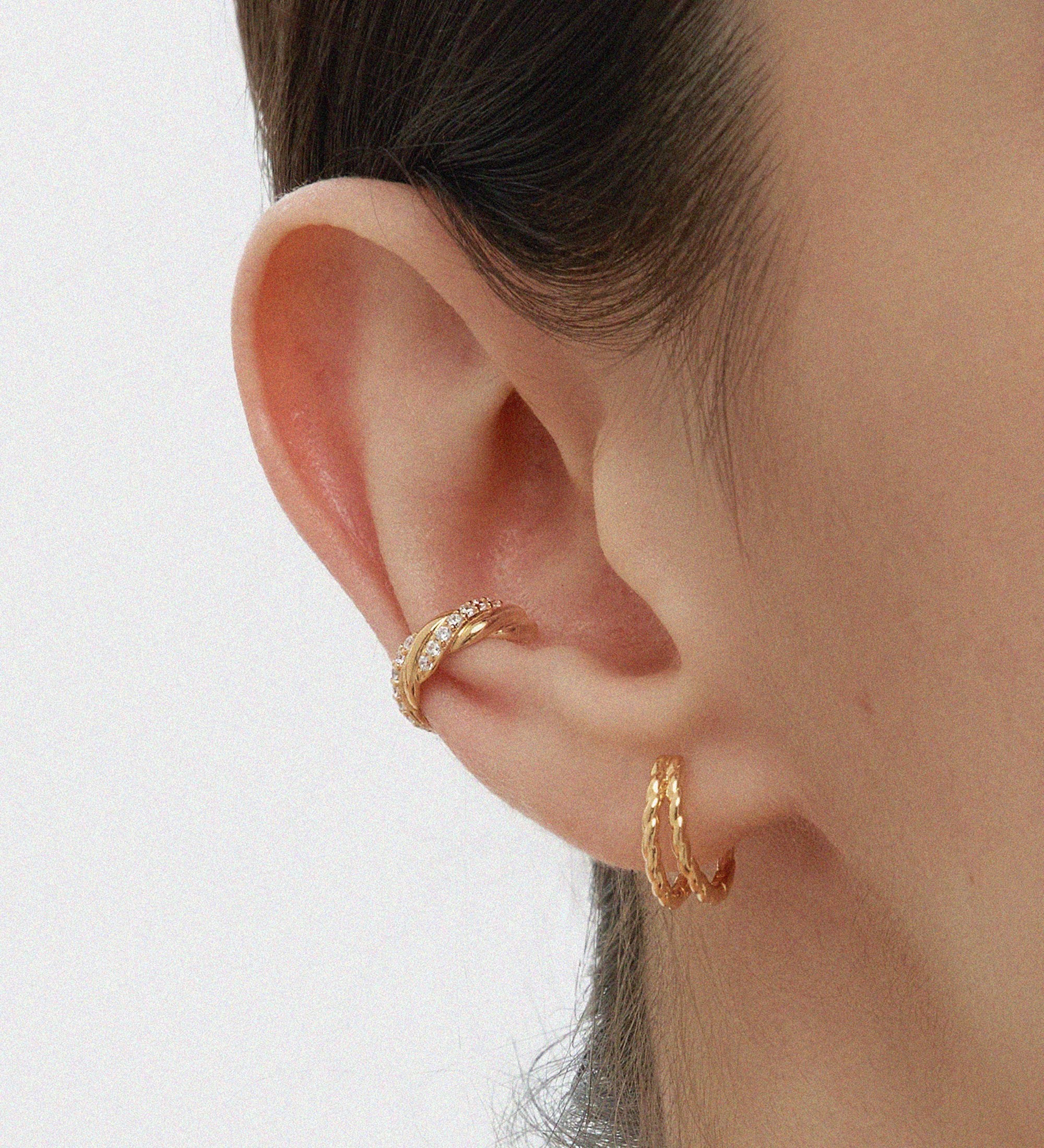 A close-up of a person’s ear wearing two gold hoop earrings and the Vera Twisted Crystal Ear Cuff in Gold, all against a plain light background.