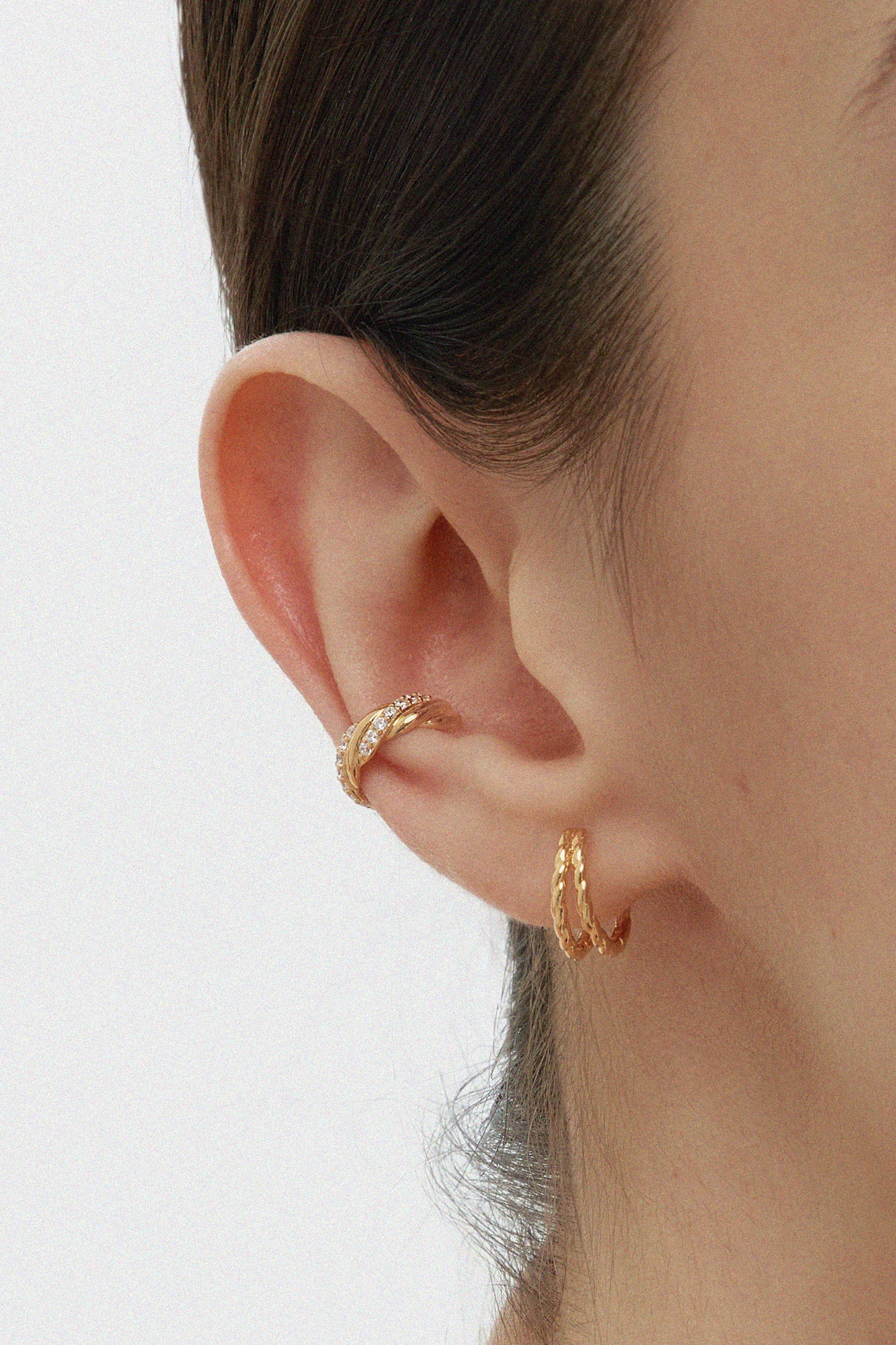 A close-up of a person’s ear wearing two gold hoop earrings and the Vera Twisted Crystal Ear Cuff in Gold, all against a plain light background.