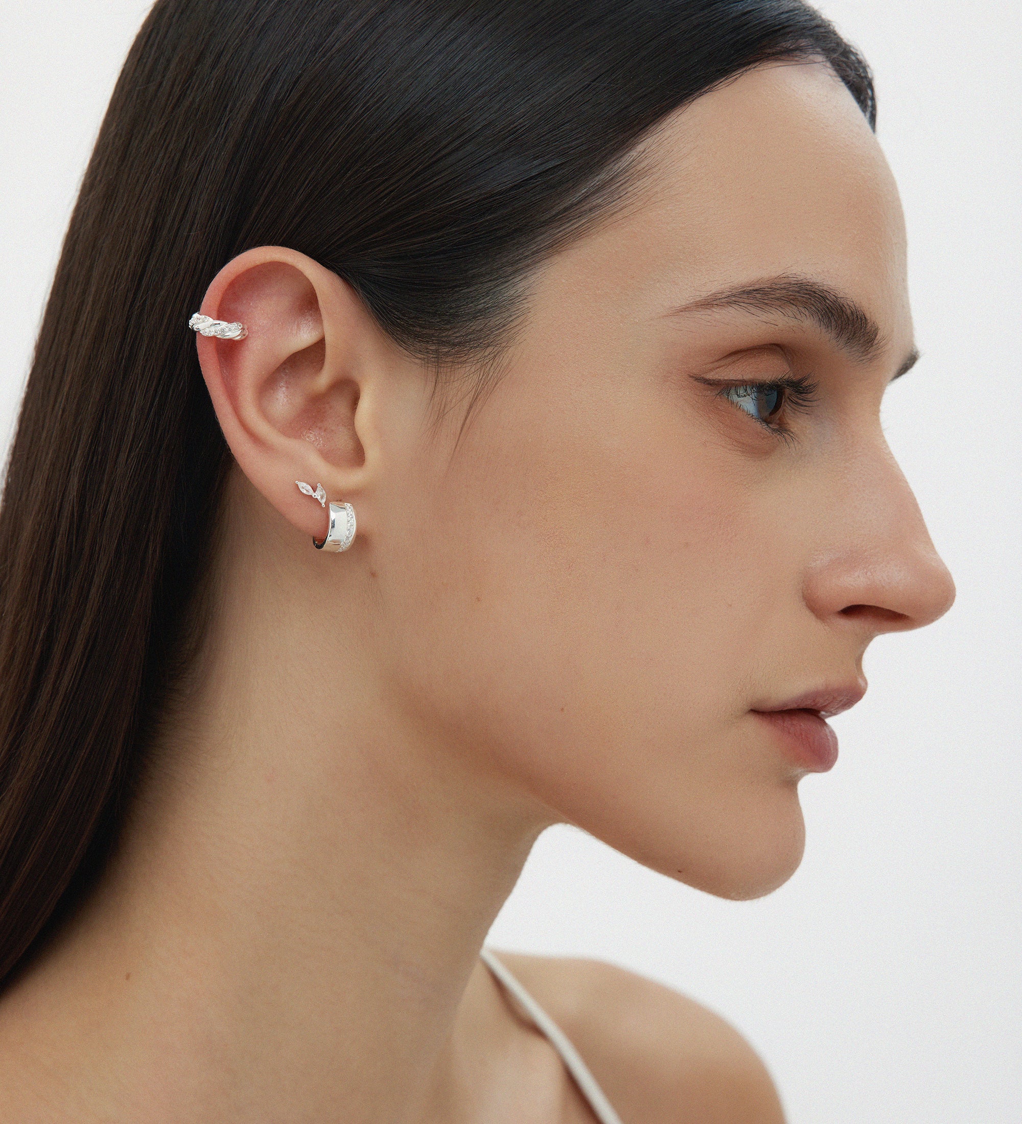 A woman with straight dark hair wears the Vera Twisted Crystal Ear Cuff in Silver on her upper ear, along with a small silver hoop in her lobe. She has natural makeup and is dressed in a white thin-strapped top.