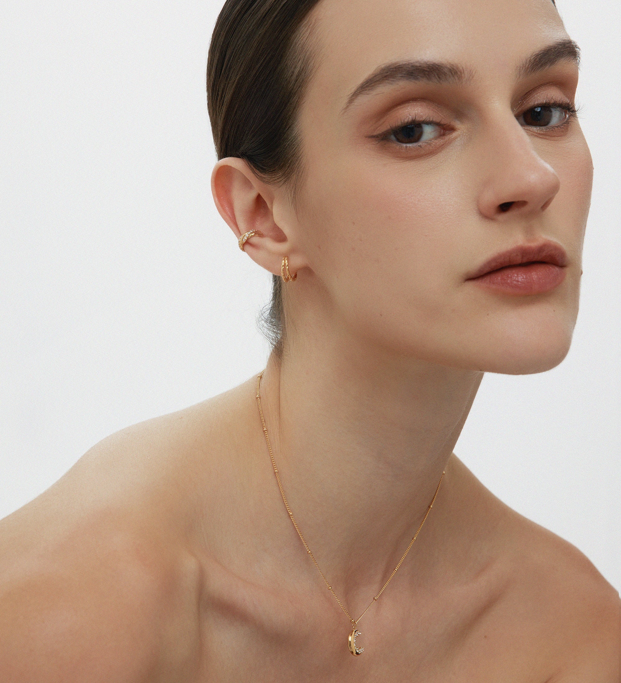 A woman with slicked-back hair wears small gold hoops, the Vera Twisted Crystal Ear Cuff in Gold, and a delicate pendant necklace, posing against a plain white background with a neutral expression.
