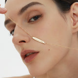 A woman with smooth skin holds the Viva La Vida Bar Chain Necklace in Gold, its adjustable figaro chain glinting as she gazes neutrally into the camera against a simple, light backdrop.