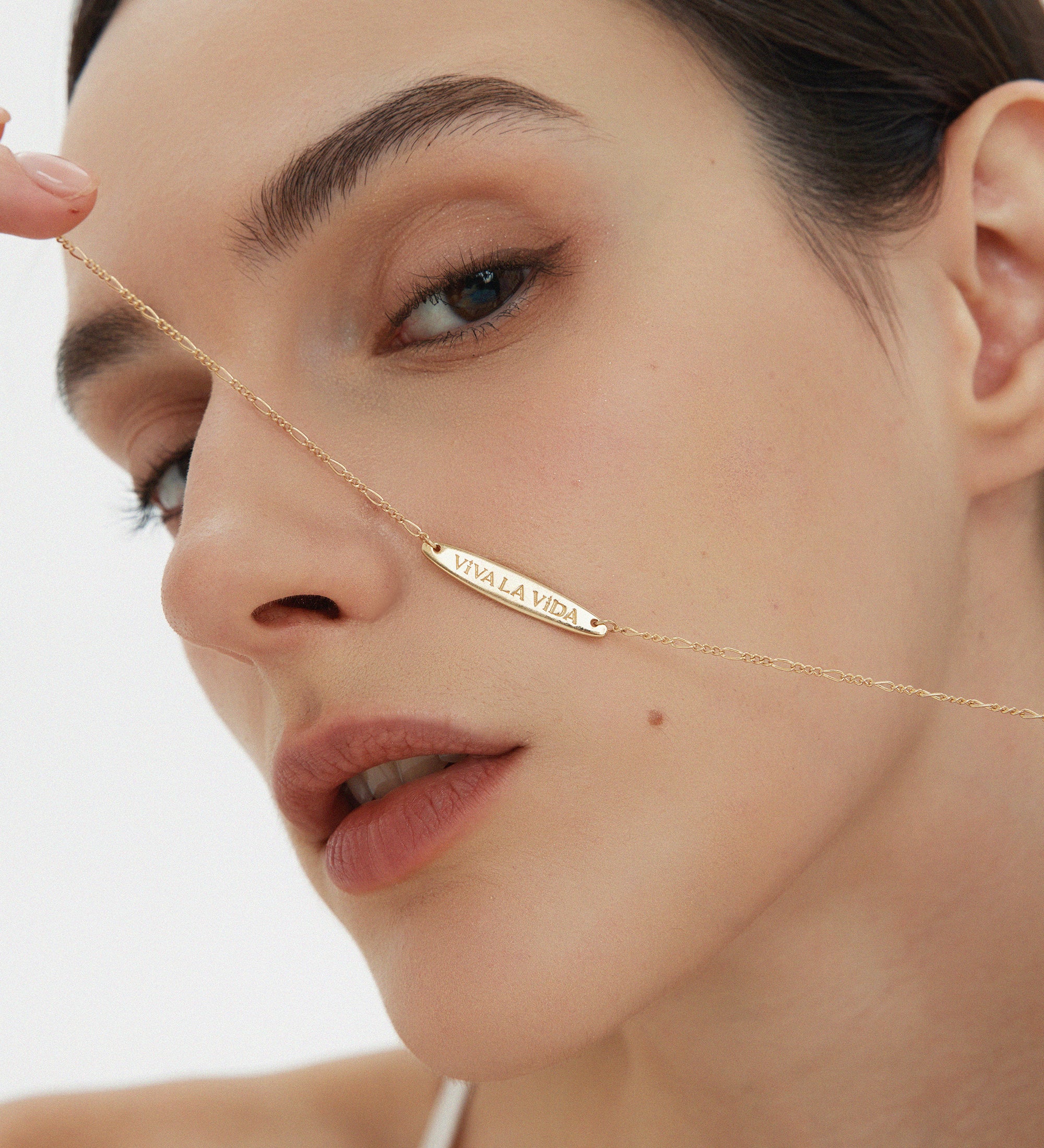 A woman with smooth skin holds the Viva La Vida Bar Chain Necklace in Gold, its adjustable figaro chain glinting as she gazes neutrally into the camera against a simple, light backdrop.