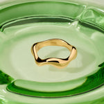 The Vica Fluid Wave Chunky Band Ring in Gold sits at the center of a smooth, modern clear green glass dish.