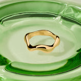 The Vica Fluid Wave Chunky Band Ring in Gold sits at the center of a smooth, modern clear green glass dish.