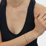 A woman in a sleeveless black top wears minimal gold jewelry—including the Vica Fluid Wave Chunky Band Ring in Gold—with her arms crossed and face partially out of frame against a plain background.