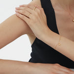 A person in a sleeveless black top rests an arm on a table, wearing the Vica Fluid Wave Chunky Band Ring in Gold along with other delicate gold jewelry. They pose against a plain white background.