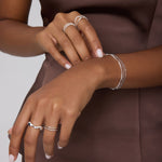 A person in a sleeveless brown satin top showcases their hands adorned with sterling silver rings and bracelets, including the Vica Fluid Wave Chunky Band Ring in Silver, against a neutral background.