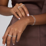 A person in a sleeveless brown satin top showcases their hands adorned with sterling silver rings and bracelets, including the Vica Fluid Wave Chunky Band Ring in Silver, against a neutral background.
