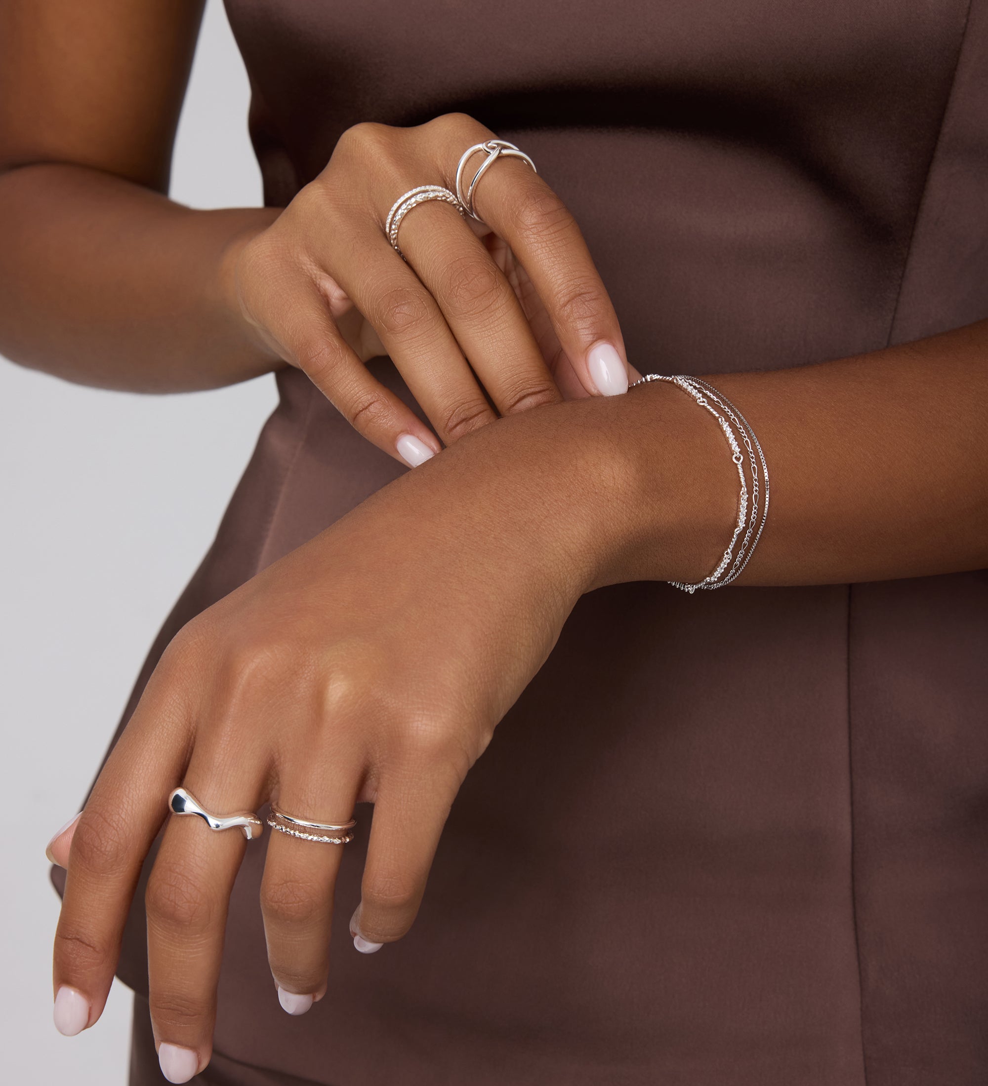 A person in a sleeveless brown satin top showcases their hands adorned with sterling silver rings and bracelets, including the Vica Fluid Wave Chunky Band Ring in Silver, against a neutral background.