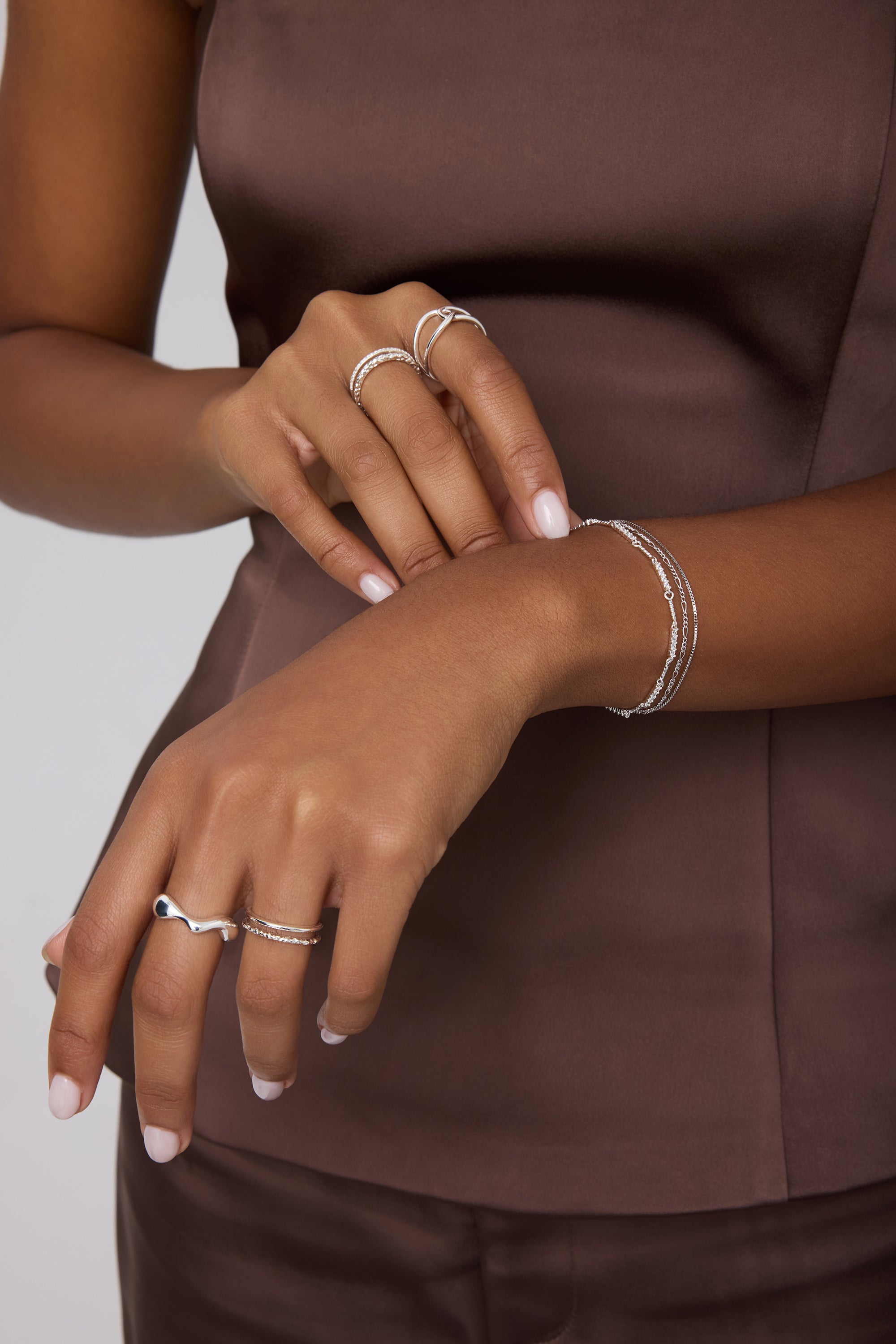 A person in a sleeveless brown satin top showcases their hands adorned with sterling silver rings and bracelets, including the Vica Fluid Wave Chunky Band Ring in Silver, against a neutral background.