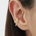 Close-up of a persons ear wearing the Vina Crystal Bezel Ear Cuff in Gold, adorned with small round cubic zirconia. Straight brown hair frames the ear, set against a softly blurred background.