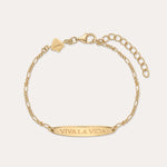 The Viva La Vida Bar Chain Bracelet in Gold features an adjustable clasp, a rectangular plate engraved with VIVA LA VIDA, and a small hexagonal charm near the clasp. Inspired by the classic Viva La Vida necklace; shown on a light beige background.