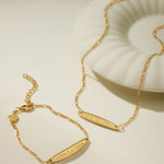 The Viva La Vida Bar Chain Bracelet in Gold, featuring an engraved bar and adjustable figaro chain, is displayed beside a white shell-shaped dish on a neutral background.