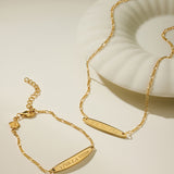 The Viva La Vida Bar Chain Bracelet in Gold, featuring an engraved bar and adjustable figaro chain, is displayed beside a white shell-shaped dish on a neutral background.