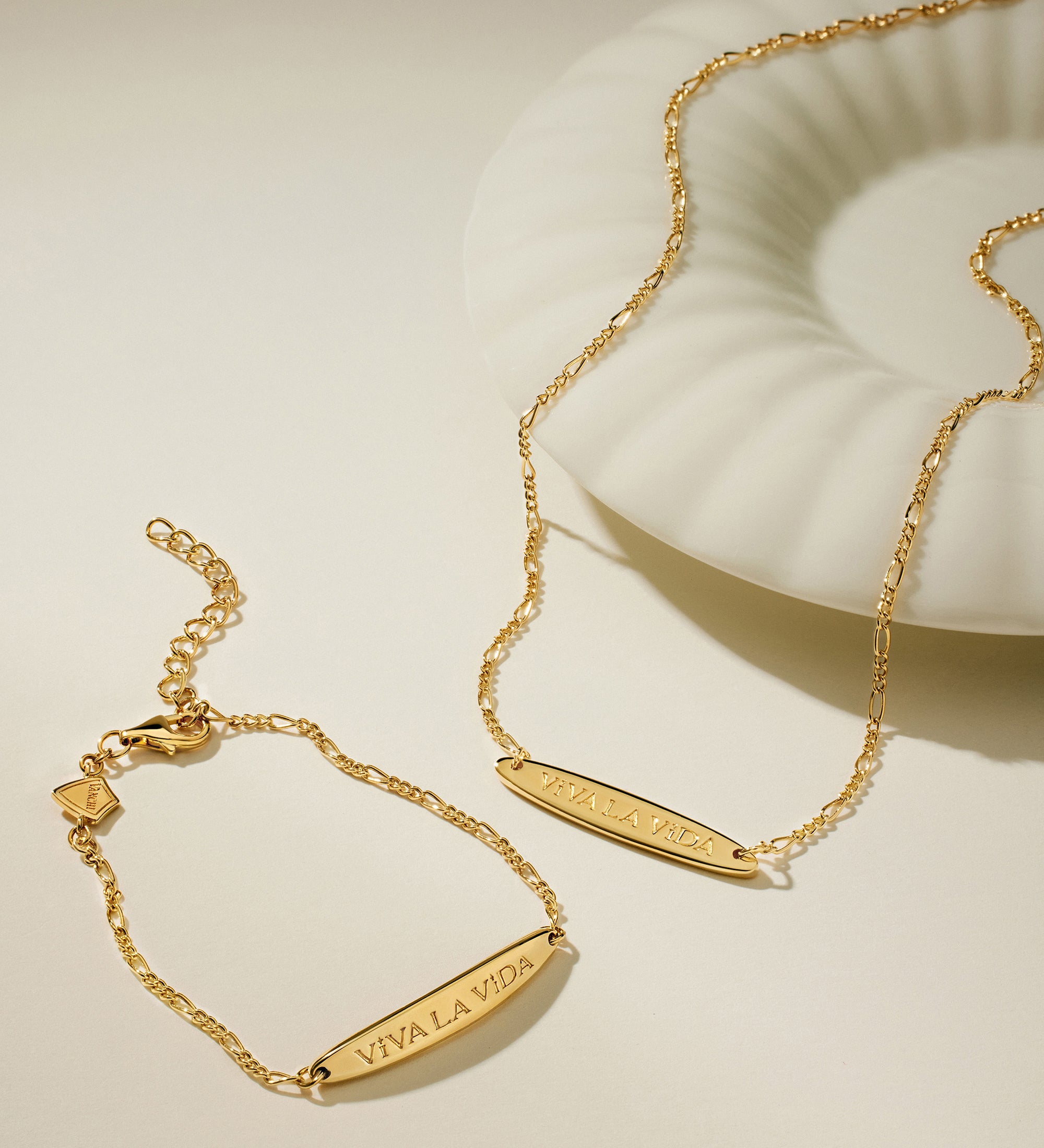 The Viva La Vida Bar Chain Bracelet in Gold, featuring an engraved bar and adjustable figaro chain, is displayed beside a white shell-shaped dish on a neutral background.