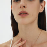 A woman with straight, dark brown hair and natural makeup wears a white thin-strapped top as she looks at the camera, holding the Viva La Vida Bar Chain Necklace in Gold—an adjustable figaro chain with an engraved bar pendant.