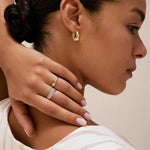 A woman with dark hair in a bun wears gold hoop earrings and the Yvette Two-Tone Mixed Metal Ring. She touches her neck with manicured fingers and is dressed in a white top featuring beige and red stripes.