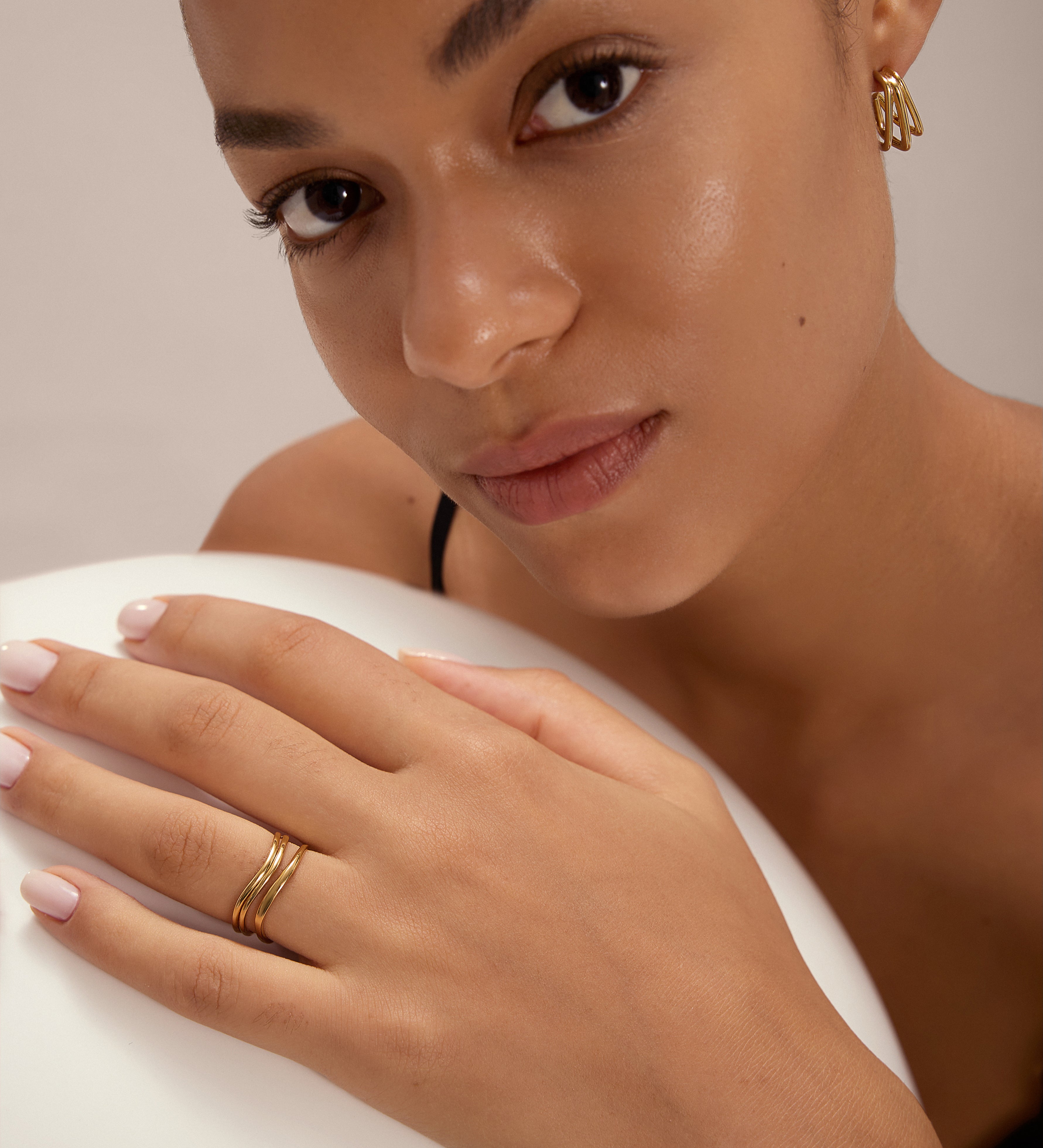 A woman with smooth skin and natural makeup wears gold plated geometric earrings and the Zoe Triple Wave Ring in Gold. She rests her manicured hand on a white surface, softly gazing at the camera.