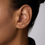 A close-up of a person’s ear showcases four gold earrings, including the 14K Recycled Solid Yellow Gold Twisted Huggie Earring in the lobe and three delicate studs along the edge. The person has medium brown skin and dark hair pulled back.