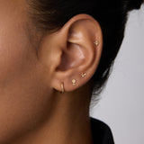 A close-up of a person’s ear showcases four gold earrings, including the 14K Recycled Solid Yellow Gold Twisted Huggie Earring in the lobe and three delicate studs along the edge. The person has medium brown skin and dark hair pulled back.