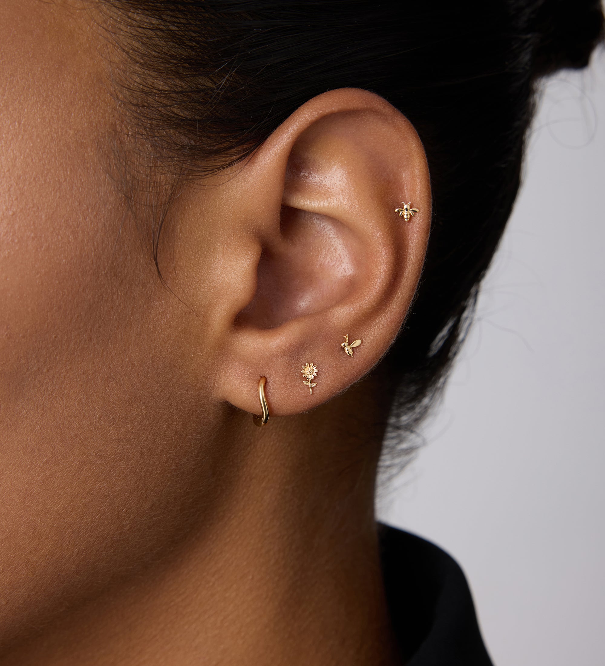 A close-up of a person’s ear showcases four gold earrings, including the 14K Recycled Solid Yellow Gold Twisted Huggie Earring in the lobe and three delicate studs along the edge. The person has medium brown skin and dark hair pulled back.