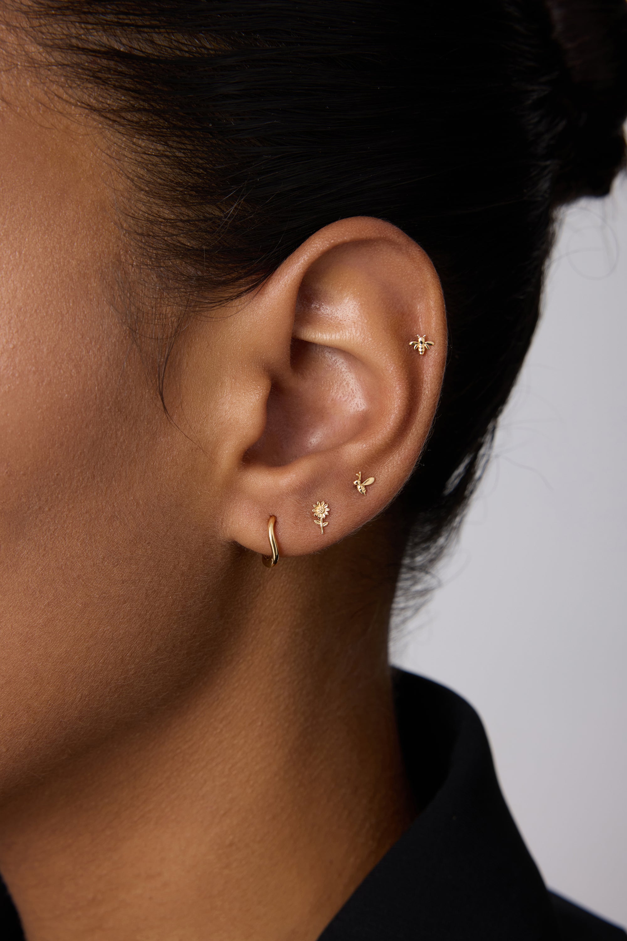 A close-up of a person’s ear showcases four gold earrings, including the 14K Recycled Solid Yellow Gold Twisted Huggie Earring in the lobe and three delicate studs along the edge. The person has medium brown skin and dark hair pulled back.